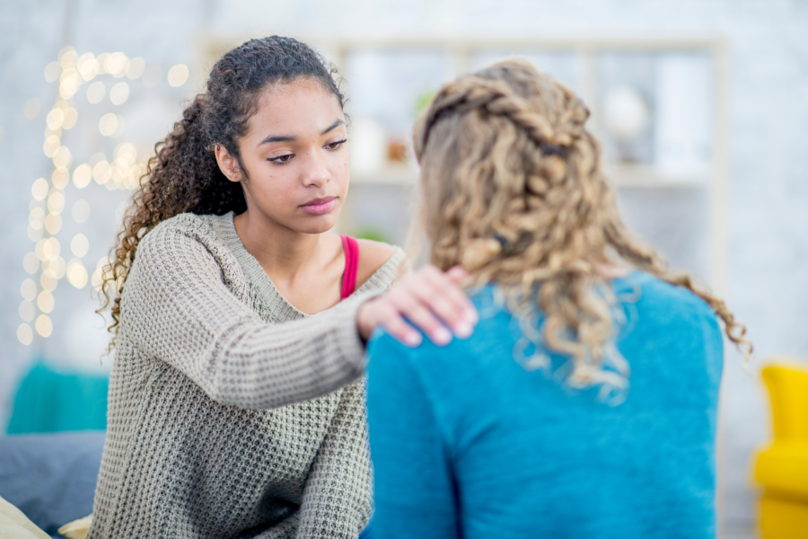 A woman with long curly brown hair putting a comforting hand on the shoulder of a woman with long blonde curly hair.